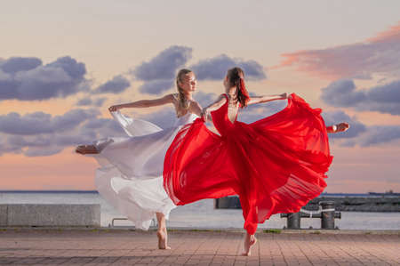 Two ballerinas in a white and red flying skirt and leotard dancing in a duet on the embankment of sea against backdrop of sunset skyの写真素材
