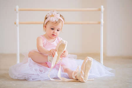 Portrait of a cute blue-eyed baby ballerina in pink sitting near the ballet barre putting on pointe shoes.の写真素材