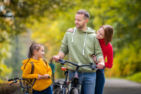 Dad, mom and daughter walk with their bicycles along the autumn path of the park.