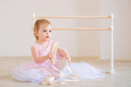 Portrait of a cute blue-eyed baby ballerina in pink sitting near the ballet barre putting on pointe shoes.の写真素材