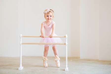 Portrait of cute blue-eyed baby ballerina in pink leotard and big pointe shoes standing near the ballet barre.の写真素材
