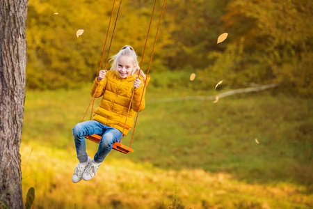Happy little blonde caucasian girl smiling and riding a rope swing in autumn in park.の写真素材