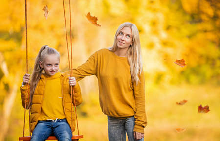 Happy young blonde mom with loose hair shakes her daughter on a swing on rope swing in autumn in park.の写真素材