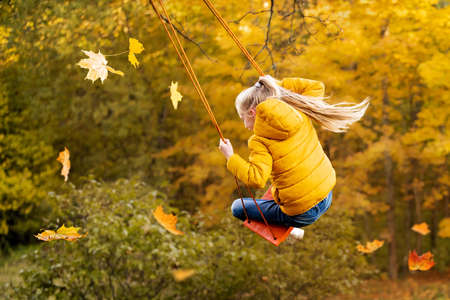 Happy little blonde caucasian girl smiling and riding a rope swing in autumn in park.の写真素材