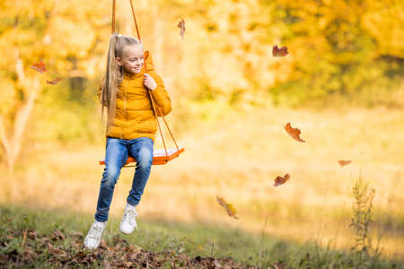 Happy little blonde caucasian girl smiling and riding a rope swing in autumn in park.の写真素材