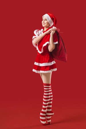 Dancing ballerina girl in pointe shoes with gift in her hands dressed as Santa Claus on red background.の写真素材