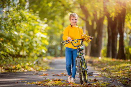 smiling girl in a yellow sweater walks with a bicycle in an autumn park on a sunny day and waves a friendly hand.の写真素材