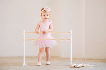 cute blue-eyed baby ballerina in a pink leotard and tutu standing near a ballet barre. Dream concept of becoming a ballerina.の写真素材