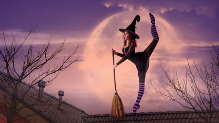ballerina on pointe shoes in black witch costume in hat and with a broom dances on roof against the backdrop of night sky and the big lilac moon on Halloween.の写真素材