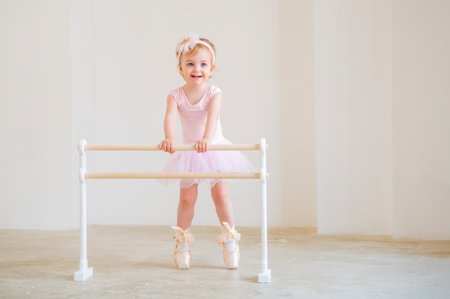 Portrait of cute blue-eyed baby ballerina in pink leotard and big pointe shoes standing near the ballet barre.の写真素材