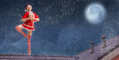 Dancing girl ballerina with gift in her hands in Santa Claus costume on roof of house on Christmas night against background of starry sky.の写真素材