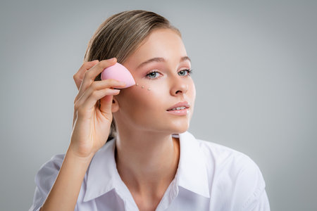 Cute beautiful natural brunette girl cleaning face with cotton sponge smiling looking at camera over white background. Cosmetology and spa.の写真素材