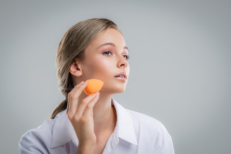 Cute beautiful natural brunette girl cleaning face with cotton sponge smiling looking at camera over white background. Cosmetology and spa.の写真素材