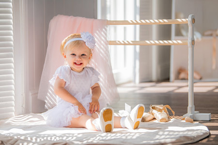 cute little ballerina in a white ballet costume is sitting near the barre in the room. Kid and ballet dance. Copyspace.の写真素材