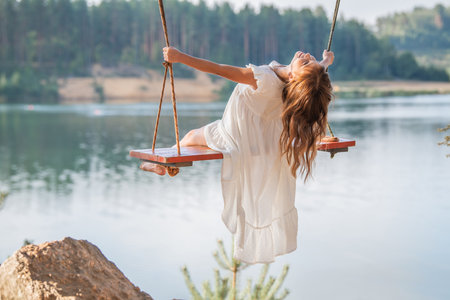girl in the forest swinging on a swing. Rope swing on a forest lake. Barefoot girl in a white dress with long hair.の写真素材