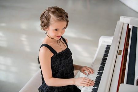 A young girl is playing the piano in a black dress. She is smiling and she is enjoying herself. The piano is white and has a black keyboardの写真素材