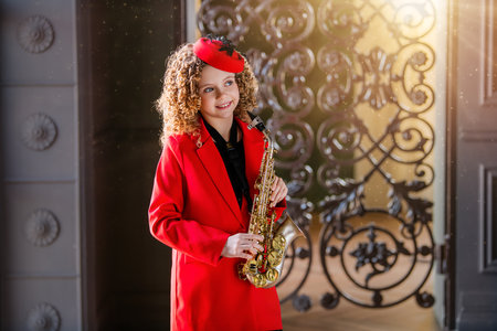A young girl in a red coat and hat is holding a saxophone. She is smiling and she is enjoying herself. Concept of joy and fun, as the girl is dressed up and playing a musical instrumentの写真素材