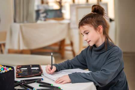 A young girl is sitting at a table with a notebook and a bag of pencils. She is smiling and holding the markers in her hands. The scene suggests that she is enjoying a creative activityの写真素材