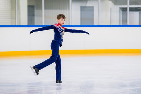 A young boy is skating on ice with his arms outstretched. He is wearing a black outfit and he is performing a trickの写真素材