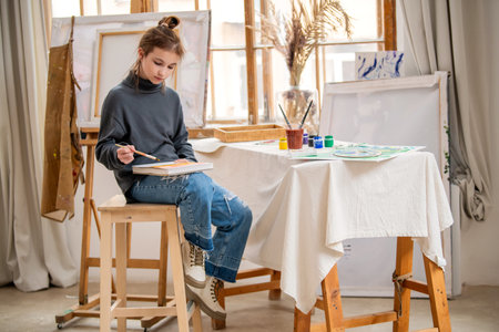 A young girl is painting a picture on a canvas. She is wearing an apron and has a brush in her hand. The painting is colorful and lively, with a mix of green and blue huesの写真素材
