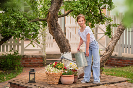 A young girl is watering plants in a garden. She is wearing a white shirt and blue jeans