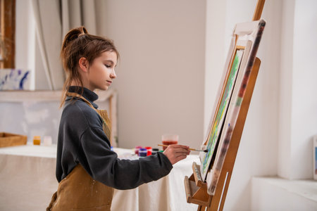 A young girl is painting a picture on a canvas. She is wearing an apron and has a brush in her hand. The painting is colorful and lively, with a mix of green and blue huesの写真素材