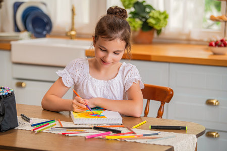 a girl draws in an album with colored pencils while sitting at a table in the interior of a country house kitchen. Development of a child's creative abilities during the holidays.の写真素材