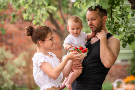A father and two children hold strawberries in a summer garden at the dacha. The scene is happy and joyfulの写真素材