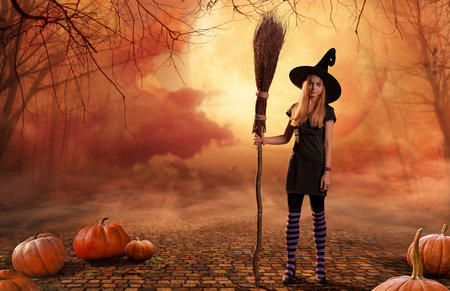 A teenage girl in a witch costume stands with a broom among pumpkins against a background of smoke and the moon on a cobblestone road.の写真素材