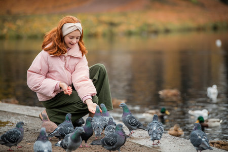 red-haired girl feeds pigeons and ducks swimming in a pond during a walk in an autumn city park. Quiet, soft, cloudy weather.の写真素材