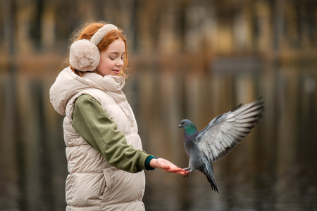 happy red-haired girl feeds pigeons from her hands during a walk in an autumn city park. Quiet, soft, cloudy weather.の写真素材