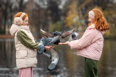 Two happy red-haired girls hand-feed pigeons during a walk in an autumn city park. Quiet, soft, cloudy weather.の写真素材