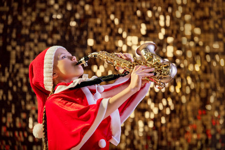 Girl child in red Christmas Santa hat playing saxophone in decorated hall. Christmas children music concept.の写真素材