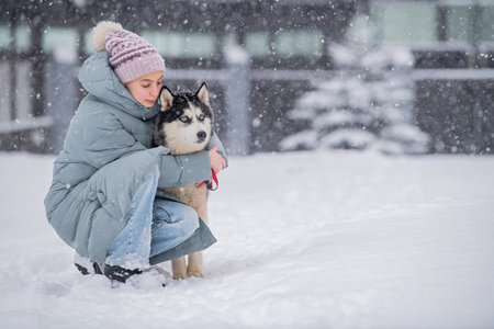 Young woman in a warm coat and beanie hugging her husky dog during a snowy winter day outdoors., dog on a walk with its ownerの写真素材