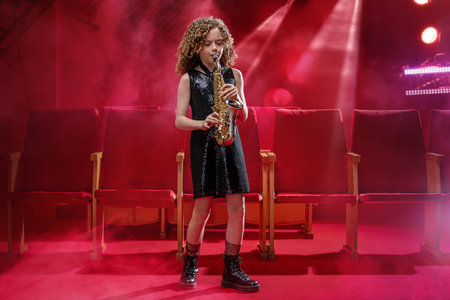 A young musician girl holds a saxophone and smiles. She is dressed in a black dress and stands against a red background in the spotlight.の写真素材