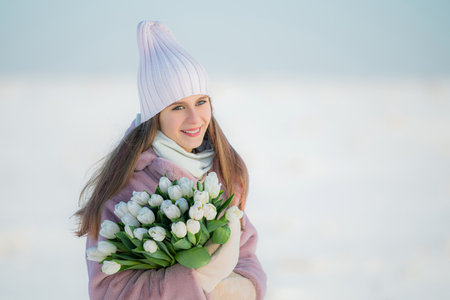 Smiling young girl in winter clothes holding bouquet of white tulips against snowy landscapeの写真素材