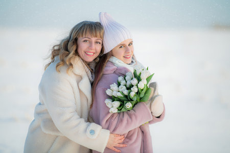Mother and Daughter in Warm Winter Coats Smiling Together in a Snowy Landscape with a Bouquet of White Tulipsの写真素材