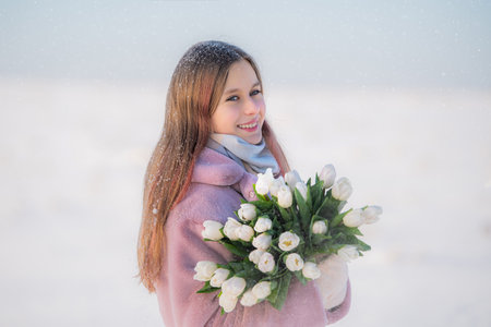 Smiling young girl in winter clothes holding bouquet of white tulips against snowy landscapeの写真素材