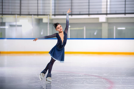 Girl figure skater in bright blue dress practicing artistic skating at indoor rinkの写真素材