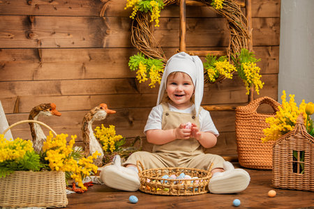 Smiling toddler in bunny hat sitting with basket of easter eggs in rural spring settingの写真素材