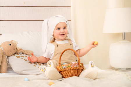 charming little girl dressed in a white bunny ears hat and beige overalls sits comfortably on a soft bed, holding an Easter egg in her hand.の写真素材