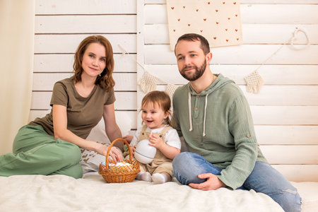 loving family of three sits on a cozy bed, enjoying Easter together. The mother, father, and their toddler share a joyful moment with a wicker basket full of colorful eggs. The warm and natural interior creates a festive and homey atmosphereの写真素材