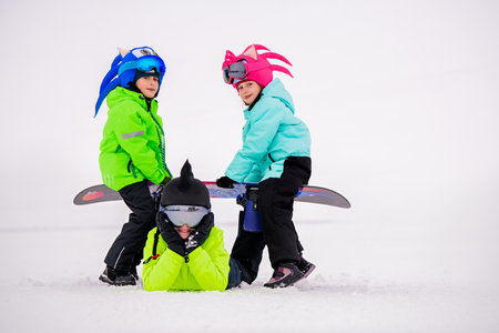 group of children in bright winter outfits play and perform tricks in the snow. In the background is a ski slope with ski lifts.の写真素材