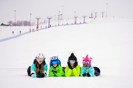 group of children and teenagers lie in the snow at a ski resort. Ski lifts and skiers are visible in the background. The atmosphere is cheerful and carefree, emphasizing the joy of winter recreation.の写真素材