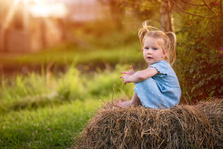 adorable little girl with pigtails in a blue dress sits barefoot on a haystack in the golden sunset light. An idyllic countryside scene filled with warmth, tranquility, and the carefree spirit of childhoodの写真素材
