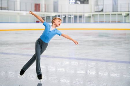 A young boy figure skater skates on the ice of an indoor ice arena and he is performing a trickの写真素材