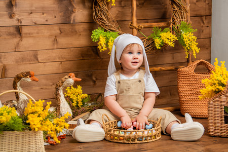 Smiling toddler in bunny hat sitting with basket of easter eggs in rural spring settingの写真素材