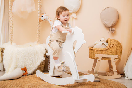 charming toddler rides a white wooden rocking horse in a beautifully decorated nursery with warm, neutral tones. The room is styled with wicker baskets, plush toys, and soft texturesの写真素材