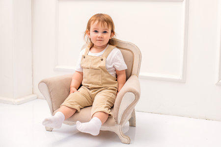cute toddler sits comfortably on a small vintage-style armchair in a bright, elegant room. The child wears beige overalls and a white t-shirt. The neutral tones and classic decor create a warm, stylish settingの写真素材