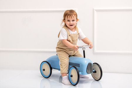 charming young child in beige overalls and a white shirt sits on a vintage blue ride-on toy car, looking curious and excited. The clean, minimalist backgroundの写真素材
